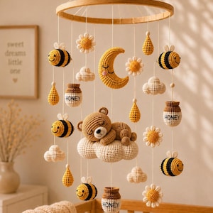 Op de afbeelding: Handgemaakte gehaakte baby mobile met een slapende beer op een wolk, bijen, bloemen, een maan en honingpotten. De mobiel hangt aan een houten ring, wat een grillige decoratie voor de kinderkamer creëert.