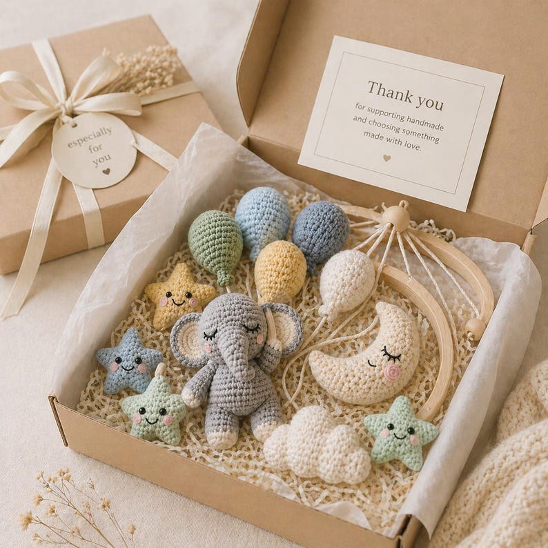 May include: A gift box containing a crochet baby mobile kit with a gray elephant, a cream moon, white clouds, and colorful balloons and stars. A card inside reads: Thank you for supporting handmade and choosing something made with love.