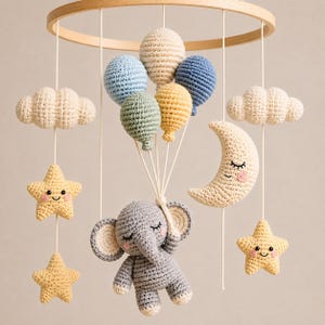 Op de afbeelding: Een gehaakte babymobiel met een grijze olifant in het midden die een bos van vijf kleurrijke ballonnen in blauw, crème, groen en geel vasthoudt. Rondom de olifant hangen twee crèmekleurige wolken, twee lachende gele sterren en een halve maan.