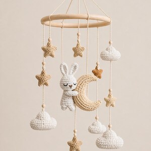 May include: A handmade baby mobile featuring a wooden ring with hanging crochet elements. The mobile includes a white bunny hugging a beige crescent moon, stars, and clouds in shades of beige and white. The mobile is suspended by natural-colored string.
