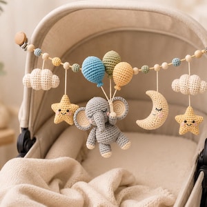 Puede incluir: Un móvil de ganchillo para cochecito con un elefante gris que sostiene tres globos en azul, verde y amarillo. El móvil incluye dos nubes color crema, dos estrellas amarillas con caras y una luna creciente con cara, ensartadas en cuentas de madera.