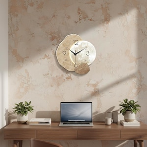 Op de afbeelding: Een decoratieve wandklok met een uniek ontwerp. De klok heeft een lichtgekleurd, onregelmatig gevormd frame met een ronde wijzerplaat. De wijzers van de klok zijn zwart. De klok is bevestigd aan een getextureerde, lichtgekleurde muur.