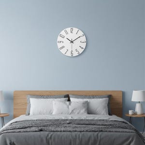 Könnte beinhalten: Eine runde Wanduhr mit weißem Zifferblatt und schwarzen Ziffern. Die Uhr verfügt über dünne schwarze Zeiger für Stunden, Minuten und Sekunden. Die Zahlen sind kreisförmig angeordnet, mit einem schlichten Design ohne sichtbaren Rahmen.