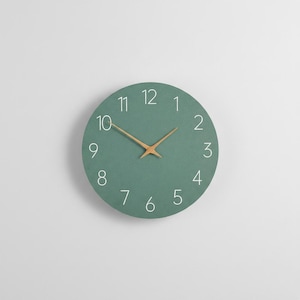 Pode incluir: Um relógio de parede redondo, verde-azulado, com algarismos brancos e ponteiros de madeira. O relógio tem um design minimalista e é adequado para a decoração da casa ou do escritório. O diâmetro do relógio é de cerca de 30 cm.