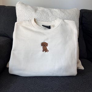 May include: A cream-colored crewneck sweatshirt featuring a small, centered embroidery of a brown curly-haired dog. The garment has long sleeves, a ribbed neckline, and a black interior brand label visible at the collar.
