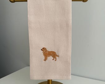 May include: A light beige waffle-weave hand towel featuring an embroidered golden retriever dog in shades of gold and brown. The towel is draped over a brass-colored metal stand with a horizontal bar and a circular base, showcasing the texture.