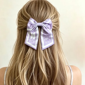 Può includere: Un fiocco per capelli in raso lavanda con strisce verticali bianche e la scritta "BTS" stampata sulle code. Il fiocco è legato al centro e viene indossato tra i capelli.