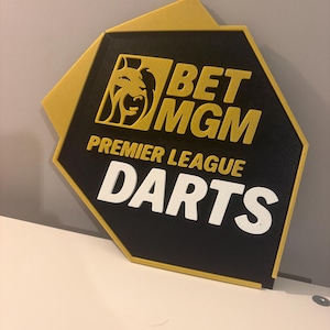 May include: A black and gold sign featuring the text "BET MGM PREMIER LEAGUE DARTS" in white and gold. The sign has a lion logo and a distinctive hexagonal shape.
