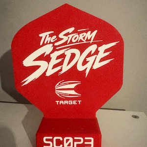 May include: A red dartboard backstop with white text reading "The Storm Sedge", "Target", and "Scop3 Steel". The backstop is uniquely shaped and designed to catch darts that miss the board, protecting the wall.