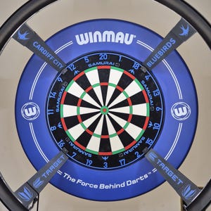 Op de afbeelding: Een blauw en zwart dartbord met de tekst "WINMAU" en "TARGET". Het dartbord heeft een cirkelvormig ontwerp met cijfers en een bullseye. De tekst "The Force Behind Darts" is ook zichtbaar. Het dartbord is omgeven door een zwart metalen frame.