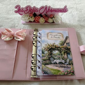 Peut inclure: Un classeur à six anneaux rose clair avec une fermeture à pression, un nœud en ruban rose et une fente pour carte. Le classeur contient un insert décoratif représentant une scène de jardin avec une table et des chaises, intitulé Mon petit coin de Paradis.