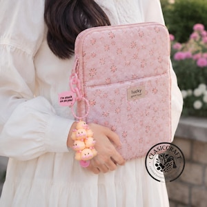 May include: A quilted pink laptop sleeve with a small floral pattern and a rectangular patch reading lucky good luck. A pink plastic chain keychain hangs from the zipper, featuring a pink speech bubble tag with the text I'm stuck on on you!