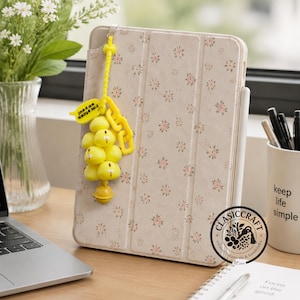 May include: A beige tablet case with a small pink and green floral pattern. A yellow keychain hangs from the side, featuring a cluster of yellow cartoon characters, a yellow chain link, and a speech bubble with the text I'm stuck on you.