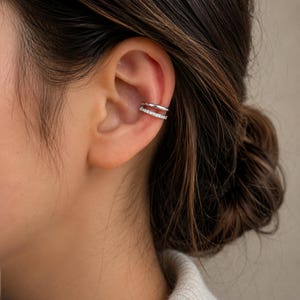 Könnte beinhalten: Ein silberfarbener Ear Cuff mit einem zweireihigen Design. Der obere Ring besteht aus glattem, poliertem Metall, während der untere Ring mit einer Reihe kleiner, klarer, funkelnder Steine besetzt ist, die das Licht reflektieren.