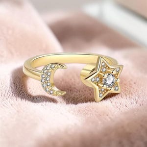 May include: A gold-colored ring featuring a crescent moon and a star design. The moon and star are embellished with small, clear gemstones. The ring is open, with the moon and star on opposite ends.