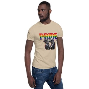 Peut inclure: T-shirt beige avec le mot "PRIDE" aux couleurs de l'arc-en-ciel. Le t-shirt présente une illustration de deux personnages qui s'embrassent. Le modèle porte un jean bleu.