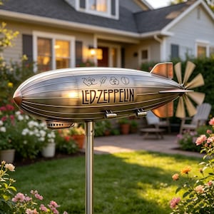 Puede incluir: Adorno de jardín metálico con forma de zepelín con el texto "LED-ZEPPELIN" y otros símbolos. El adorno tiene una hélice y está montado sobre un poste plateado. El fondo incluye una casa, hierba verde y flores.