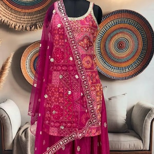 Puede incluir: Un vibrante conjunto étnico de color fucsia exhibido en un maniquí. El conjunto incluye una parte superior estampada, una falda a juego y una dupatta transparente. El atuendo está adornado con intrincados bordados y adornos.