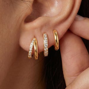May include: Three gold hoop earrings worn in a single earlobe. The first hoop is a double-band design with clear stones, the second is a single band with clear stones, and the third is a plain, smooth gold hoop. All earrings have a polished finish.