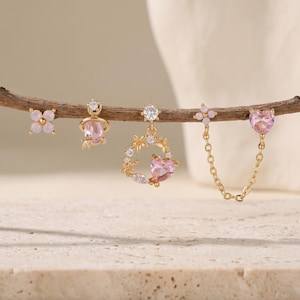 May include: A set of five gold-tone earrings featuring light pink stones and clear crystals. The designs include a four-petal flower, a planet shape, a circular floral wreath with a heart, and a double-stud earring connected by a gold chain.