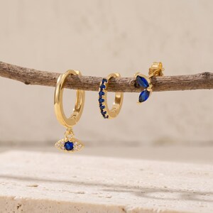 Pode incluir: Três brincos dourados exibidos num ramo fino. Um é uma argola com um pendente em forma de olho com uma pedra central azul e detalhes claros. Outro é uma argola pequena com pedras azuis. O terceiro é um brinco de pino com pedras azuis.
