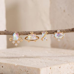 May include: Four gold-tone stud earrings featuring iridescent oval and rectangular stones. The designs include a fleur-de-lis shape, a planet with a ring, a twisted bar, and a starburst setting, all accented with small clear round stones.