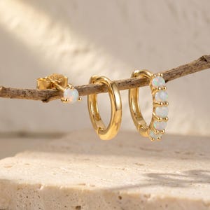 May include: A set of three gold-toned earrings consisting of one stud earring with a single round opal, one plain gold hoop earring, and one gold hoop earring featuring four small round opals set along the outer edge of the circular band.