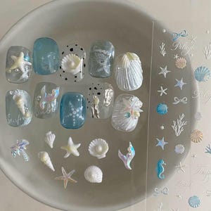 Op de afbeelding: Een set nageldecoraties met diverse 3D-bedels in zeethema, waaronder zeesterren, schelpen en zeemeerminstaarten in blauwe, witte en parelmoerkleurige tinten, inclusief een transparant vel met bijpassende nautische stickers.