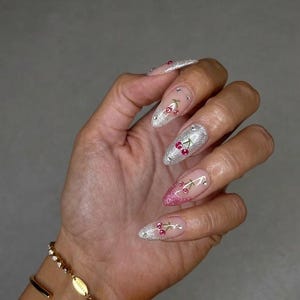 May include: A hand displaying long, almond-shaped nails. The nails are decorated with silver glitter, cherry designs, and small rhinestones. Some nails have a pink glitter gradient. A gold bracelet with a chain and oval charm is on the wrist.