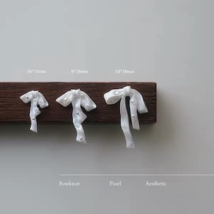 Op de afbeelding: Drie witte strikvormige bedels met ingebedde kleine parels, weergegeven op een donkere houten strip. De maten zijn gelabeld als 10*14mm, 9*18mm en 13*19mm. De tekst Bowknot, Pearl, Aesthetic staat onder de bedels op een lichte achtergrond.