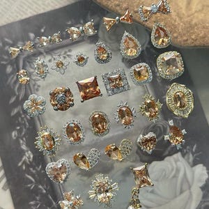 Könnte beinhalten: Eine Sammlung verschiedener dekorativer Anhänger mit champagnerfarbenen Kristallen in silberfarbenem Metall. Die Stücke haben verschiedene Formen wie Schleifen, Herzen, Blumen, Ovale, Quadrate und Tropfen, einige mit umgebenden klaren Strasssteinen.