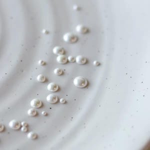 Könnte beinhalten: Eine Ansammlung kleiner, glatter, weißer, kugelförmiger Perlen, die auf einer hell cremefarbenen Oberfläche verstreut sind. Die Oberfläche weist eine subtile, wellenförmige Textur mit feinen dunklen Sprenkeln auf.