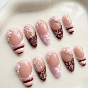 Op de afbeelding: Een set van tien kunstnagels met een lichtroze basis en diverse ontwerpen. Sommige nagels hebben witte bloempatronen, terwijl andere bruine uiteinden hebben met witte krullen. Accentueren met parels en een ster.