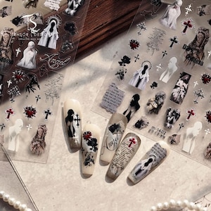 May include: Nail art stickers with gothic and religious themes. Designs include angels, crosses, and script, in black, white, and red. The word "Angel" is visible on one sticker. The stickers are on a clear sheet.