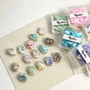 Könnte beinhalten: Eine Sammlung ovaler Nagelkunst-Tipps mit irisierenden, perlmuttartigen und marmorierten Oberflächen in Pastellfarben, akzentuiert mit kleinen goldfarbenen Ketten, Perlen und Sternen. Mehrere klare quadratische Behälter mit der Aufschrift Rimuru enthalten passende Flocken.
