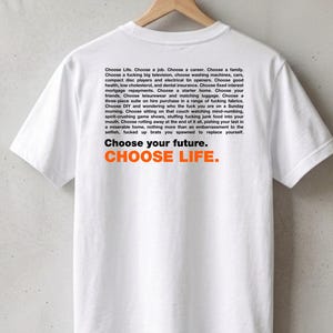 Pode incluir: Uma t-shirt branca de manga curta com um bloco de texto preto que diz: Choose Life. Choose a job. Choose a career. Choose a family. Choose a fucking big television... Choose your future. CHOOSE LIFE. em letras pretas e laranjas a negrito.