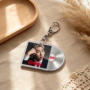May include: Silver-toned keychain featuring a rectangular image of a person in a suit and a red bow tie, with a silver record-shaped design. The keychain has a silver clasp for attaching to keys or bags.