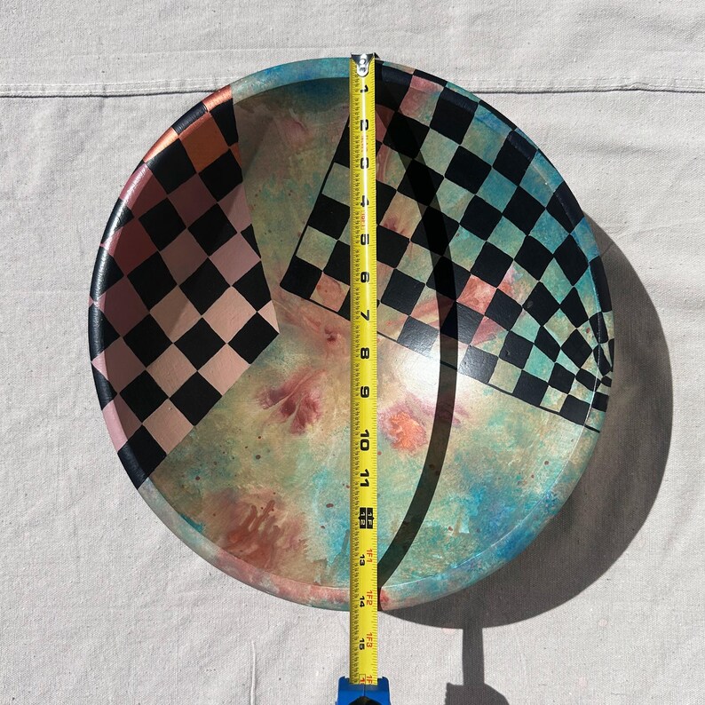 K&ouml;nnte beinhalten: Eine runde dekorative Sch&uuml;ssel mit einem farbenfrohen, abstrakten Design. Die Sch&uuml;ssel hat ein Schachbrettmuster in Schwarz und Rosa sowie Schwarz und Blaugr&uuml;n. Die Sch&uuml;ssel hat einen Durchmesser von etwa 38 cm.