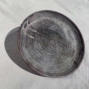 Könnte beinhalten: Ein handgefertigter Keramikteller mit einer dunkelgrauen und weißen, gesprenkelten Oberfläche. Der Teller hat eine kreisförmige Form mit einem leicht erhöhten Rand und einem Durchmesser von etwa 20 cm. Das Design des Tellers zeigt konzentrische Kreise.