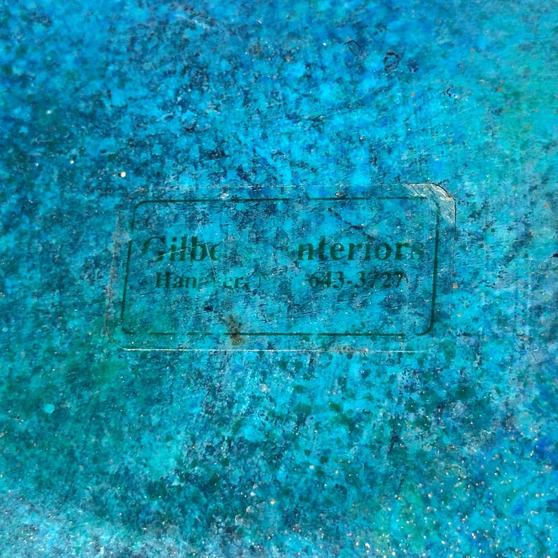 K&ouml;nnte beinhalten: Nahaufnahme einer strukturierten Oberfl&auml;che in T&uuml;rkis- und Blaut&ouml;nen. Ein rechteckiges Etikett mit dem Text "Gilbe Interiors" und einer Telefonnummer ist sichtbar. Die Oberfl&auml;che hat ein gesprenkeltes Aussehen.