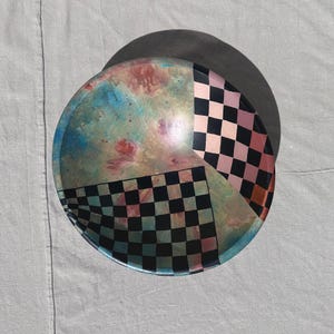 K&ouml;nnte beinhalten: Eine runde dekorative Schale mit einem einzigartigen Design. Die Schale hat ein farbblockartiges Muster mit Abschnitten aus schwarz-wei&szlig;em Schachbrettmuster und einem Abschnitt mit einer Mischung aus Blau-, Rosa- und Goldt&ouml;nen. Die Schale steht vor einem neutralen Hintergrund.