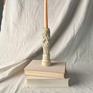 May include: A tall, off-white candle holder featuring a sculpted relief of a figure with flowing hair and intricate details, resting on a circular base with a geometric pattern. A single, slender, peach-colored taper candle is inserted into the top.