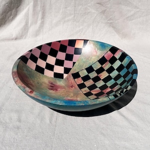 K&ouml;nnte beinhalten: Oval geformte Dekoschale mit einem farbenfrohen, abstrakten Design. Die Schale hat ein Schachbrettmuster in Rosa und Schwarz sowie ein weiteres in Schwarz und Blau. Die Au&szlig;enseite ist t&uuml;rkis und die Innenseite hat einen Marmoreffekt.