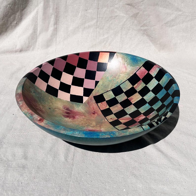 K&ouml;nnte beinhalten: Oval geformte Dekoschale mit einem farbenfrohen, abstrakten Design. Die Schale hat ein Schachbrettmuster in Rosa und Schwarz sowie ein weiteres in Schwarz und Blau. Die Au&szlig;enseite ist t&uuml;rkis und die Innenseite hat einen Marmoreffekt.