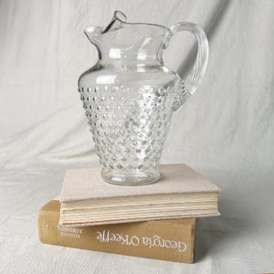 Puede incluir: Una jarra de vidrio transparente con un patrón de puntos en relieve en el cuerpo, un asa curva y un pico vertedor ancho. La jarra está colocada sobre dos libros apilados, uno con cubierta beige y otro con cubierta color canela con el título Georgia O'Keeffe.