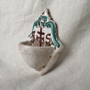 Może przedstawiać: Ścienna kropielnica ceramiczna z kremowym szkliwem i widocznym wykończeniem typu krakle. Na tylnej płycie znajduje się ręcznie malowany brązowy krzyż z literami JHS oraz ozdobne zielone akcenty w kształcie wolut wzdłuż górnej krawędzi.