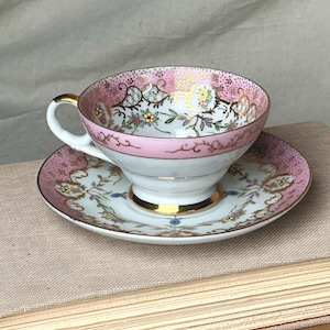 May include: A porcelain teacup and saucer set featuring a white base with a pink rim, gold gilded accents, and a delicate floral pattern. The interior of the cup and the edge of the saucer are decorated with intricate gold filigree and small flowers.