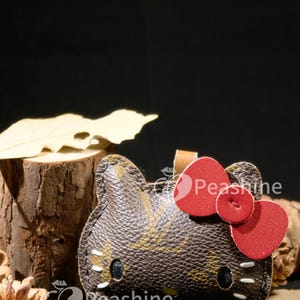 May include: A brown and tan leather keychain shaped like a cartoon cat's face, with a red bow. The keychain has black eyes and white whiskers. The word "Peashine" is printed on the keychain.