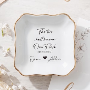 May include: A square white ceramic trinket dish with a scalloped gold-trimmed edge. The center features black text reading The two shall become One Flesh, Ephesians 5:31, and the names Emma and Allen separated by a small black heart symbol.