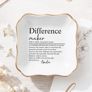 May include: A white ceramic trinket dish with a gold rim, inscribed with the definition of "Difference maker" in black. The definition includes the name "Amelia". Flowers and cards are in the background.